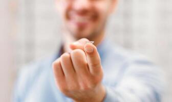 A man's hand holds a contact lens. He is smiling and out of focus in the background. His hand is outstretched, holding the contact lens in the middle of his index finger. photo