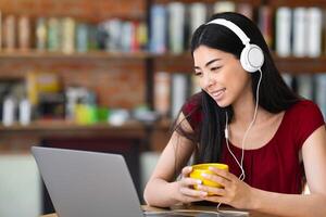 Smiling Asian Woman Watching Webinar With Laptop And Headphones In Cafe And Drinking Coffee, Having Conference Or Online Lesson, Looking AT Computer Screen And Smiling, Enjoying E-Learning photo