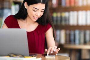 Useful App. Cheerful Asian Woman Browsing New Application On Smartphone In Cafe While Working Remotely With Laptop Computer photo