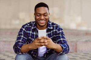 Lifestyle Concept. Happy afro man sitting in the city, listening to music and typing text message on smartphone device photo