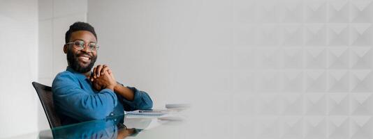 A young African American entrepreneur sits confidently at his desk, managing daily tasks in a vibrant office. He smiles, embodying success and motivation in a dynamic work setting, copy space photo