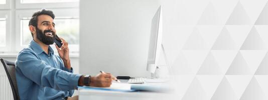 Positive thoughtful indian businessman talking on cellphone and taking notes while working on computer in office. Mobile business communication, successful entrepreneurship career concept photo