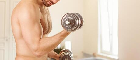 Unrecognizable Shirtless Muscular Man Exercising With Dumbbells Training Standing At Home, Pumping Biceps Muscles. Domestic Male Workout With Dumbbell. Sporty Lifestyle. Cropped, Panorama, Free Space photo
