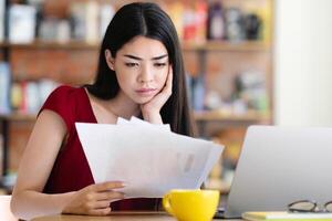 Concentrated asian female employee working with papers and laptop at cafe, having problem, checking documents while sitting at table, copy space photo