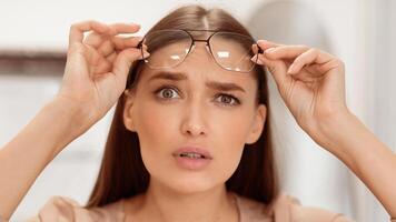 The woman appears puzzled as she holds her eyeglasses close to her face. Her expression reflects confusion, possibly about something she is viewing. The indoor setting is softly lit. photo