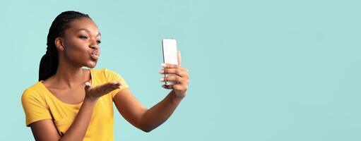 A woman in a yellow shirt playfully blows a kiss towards her smartphone while taking a selfie. The background is a vibrant teal, adding to the cheerful atmosphere of the moment. photo