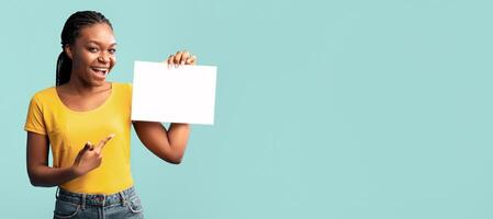 A woman with braids smiles enthusiastically while pointing at a blank piece of paper. She stands against a pastel-colored background, radiating positivity and energy. photo