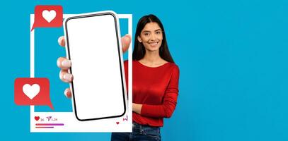 An influencer interacts with her smartphone against a vibrant blue backdrop, highlighting a blank screen for social media use. She presents chatting features depicted with hearts and comments. photo