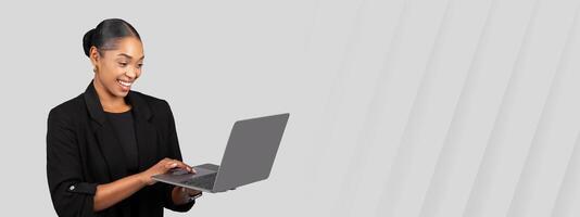Focused smiling African American businesswoman working on a laptop with a pleased expression, signifying productivity and digital engagement in a sleek black suit on grey background, studio photo