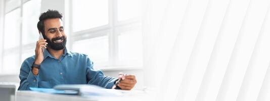 Smiling indian businessman talking on cellphone and noting down information, man entrepreneur speaking on mobile phone and taking notes, sitting at desk in office photo