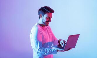 A young man stands with a laptop, engaged in his work. The background is illuminated with vibrant pink and blue hues, creating a dynamic and modern atmosphere. photo