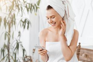 Happy girl checking on social media while applying cream on face after shower, using smartphone, empty space photo