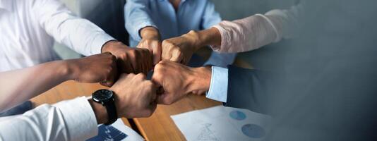 Teamwork Concept. Multiethnic group of business people making fist bumps together while sitting at desk in modern office, company employees joining hands as sign of unity and success, closeup photo