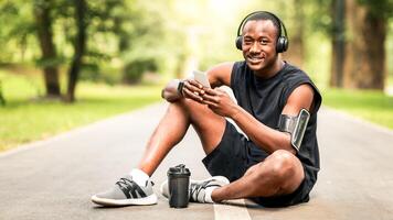Joyful african sportsman resting on jogging trail at park, using phone and drinking water, empty space photo
