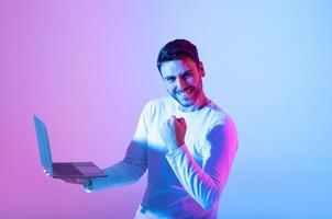 A young man in a light sweater expresses joy and triumph while using a laptop. The colorful background features shades of blue and pink, enhancing the celebratory atmosphere. photo