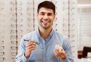 A man stands in an eyewear store showcasing a pair of glasses and a contact lens case, smiling. The background features a display of various eyeglasses, highlighting a friendly atmosphere. photo