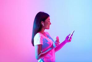 A young woman with long hair holds a smartphone and a wireless earbud, smiling as she listens to music. The colorful background features shades of pink and blue, enhancing her enjoyment. photo
