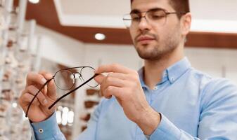 A man in a light blue shirt inspects a pair of glasses in an optical shop. He thoughtfully evaluates their design and fit, surrounded by various eyewear options on display. photo