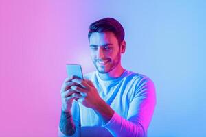 A young man enjoys using his smartphone, smiling as he interacts with it. The background features vibrant blue and pink lighting, creating a modern, energetic atmosphere. photo