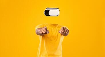 A person is immersed in the world of virtual reality, wearing goggles without a visible head or face. They point forward enthusiastically against a bright yellow background, showcasing modern tech. photo