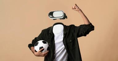 A person without a visible head or face is immersed in virtual reality, wearing goggles and holding a soccer ball. The setting features a plain background, highlighting the modern technology. photo