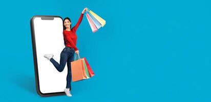 A cheerful customer holds shopping bags while stepping joyfully from a large smartphone screen. She embodies the excitement of online shopping and modern commerce in a vibrant setting. photo