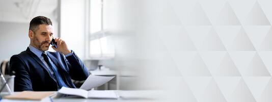 A middle aged businessman engaged in a phone call while reviewing documents in a contemporary office. He appears focused and professional, embodying entrepreneurial success and ambition, copy space photo