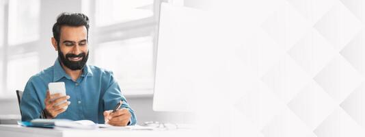 Cheerful indian bearded man in blue shirt enjoys text on his phone as he takes notes with a pen in a bright, modern office setting photo