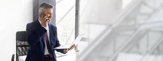 Middle Aged Businessman In Suit Talking On Cellphone And Using Laptop At Office, Mature Boss Looking At Computer Screen And Having Business Call With Client While Leaning At Desk, Copy Space photo