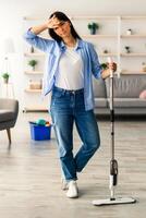 A young woman stands in her living room with a mop, looking tired after a long cleaning session. Furniture is neatly arranged, showcasing a cozy home atmosphere. photo