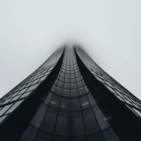 Modern skyscraper, shrouded in mist. Vast, glass-paneled structure, viewed from below, reaching towards a hazy sky photo