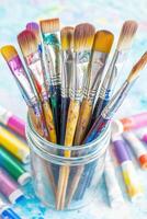 Well-used artist paintbrushes with dried paint in a glass jar and paint tubes displayed on a colorful surface, creating an artistic and creative environment photo