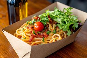 Spaghetti with bacon, cherry tomatoes and arugula served in a cardboard container photo