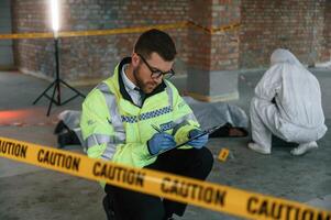 Man, in glasses is holding notepad and pen. Detectives are collecting evidence in a crime scene near dead body photo