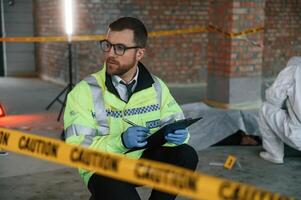 Man in glasses is holding notepad and pen. Detectives are collecting evidence in a crime scene near dead body photo