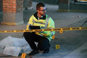 Male detective is collecting, evidence, in a crime scene near dead body on the construction site. Sitting with notepad in hands photo