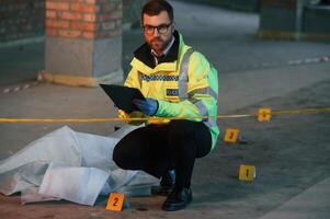 Male detective is collecting evidence in a crime scene ,near dead body on the construction site. Sitting with notepad in hands photo