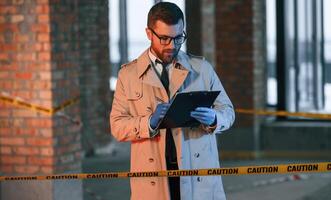 Male detective in coat is collecting evidence in a crime, scene on the construction site. With notepad in hands photo