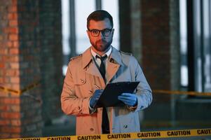 Male detective i,n coat is collecting evidence in a crime scene on the construction site. With notepad in hands photo