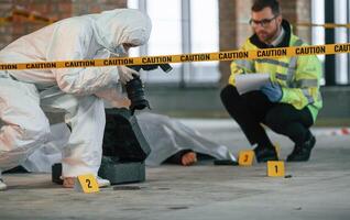 Woman is making photos by camera. Detectives are collecting evidence in a crime scene near dead body