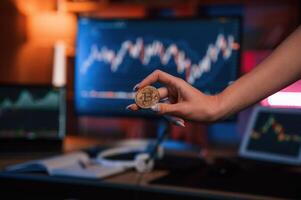 Close, up view of woman's hand that holding bitcoin against computer display and tablet with graphs photo