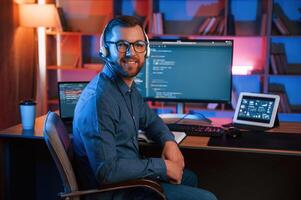Positive professional programmer is working, indoors and smiling. Neon lighting photo