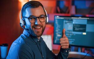Showing thumb up. Professional, programmer, is working indoors. Neon lighting photo