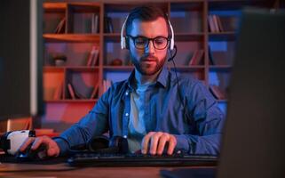 Front, view. Man is sitting by a computer indoors at home. Neon lighting photo
