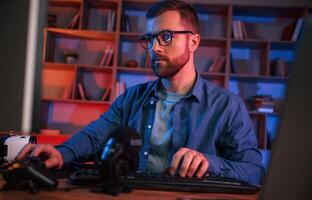 Front view. Man is sitting by a computer indoors at, home. Neon lighting photo