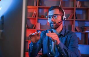 Having online meeting. Man is sitting by a computer, indoors at home. Neon lighting photo
