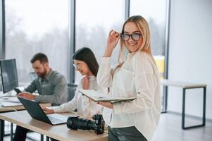Woman in glasses is standing ,with notepad in hands. Successful team are working together in the office photo