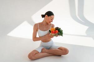 Looking at healthy food. Sitting on the floor. Young caucasian woman with slim body shape is indoors in the studio photo