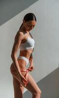 Using, the, measuring tape. Young caucasian woman with slim body shape is indoors in the studio photo