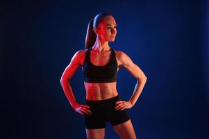 Standing, with the hands ,on waist. Beautiful muscular woman is indoors in the studio with neon lighting photo